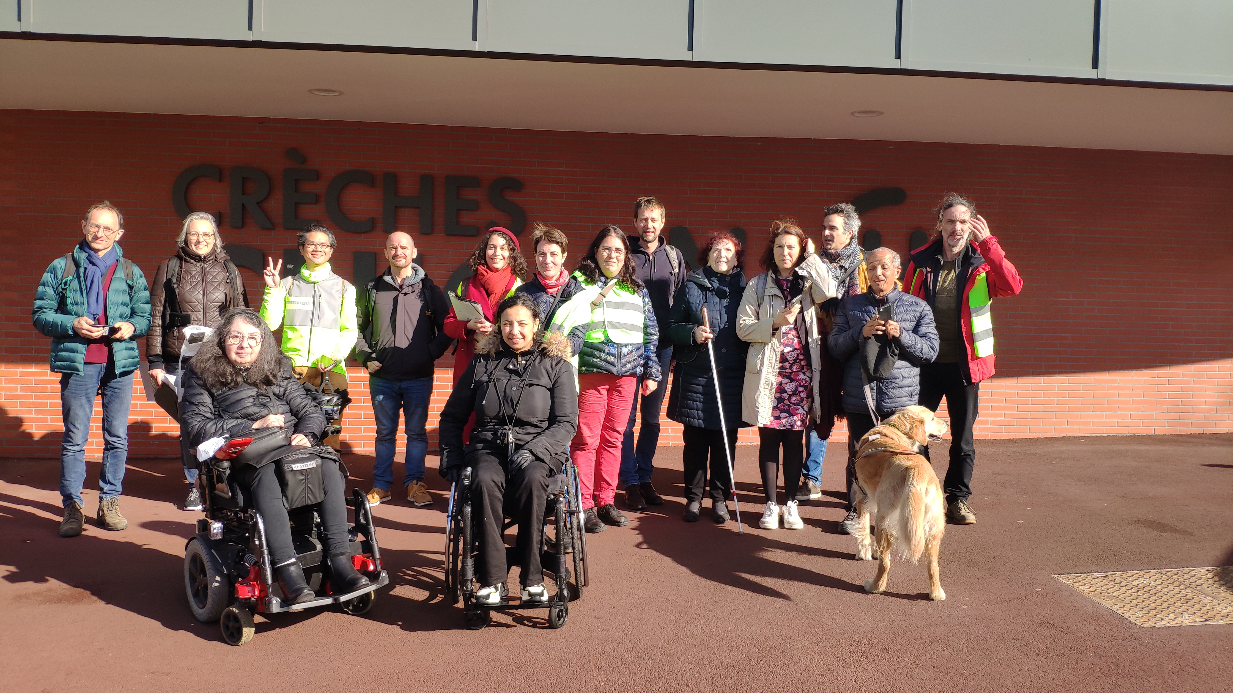 Un groupe d'une douzaine de personnes (personnes  valides et en situation de handicap, en fauteuil, avec chien guide, certains avec des chasubles fluos) prend la pose au soleil pour une photo, devant un bâtiment moderne de Créteil.