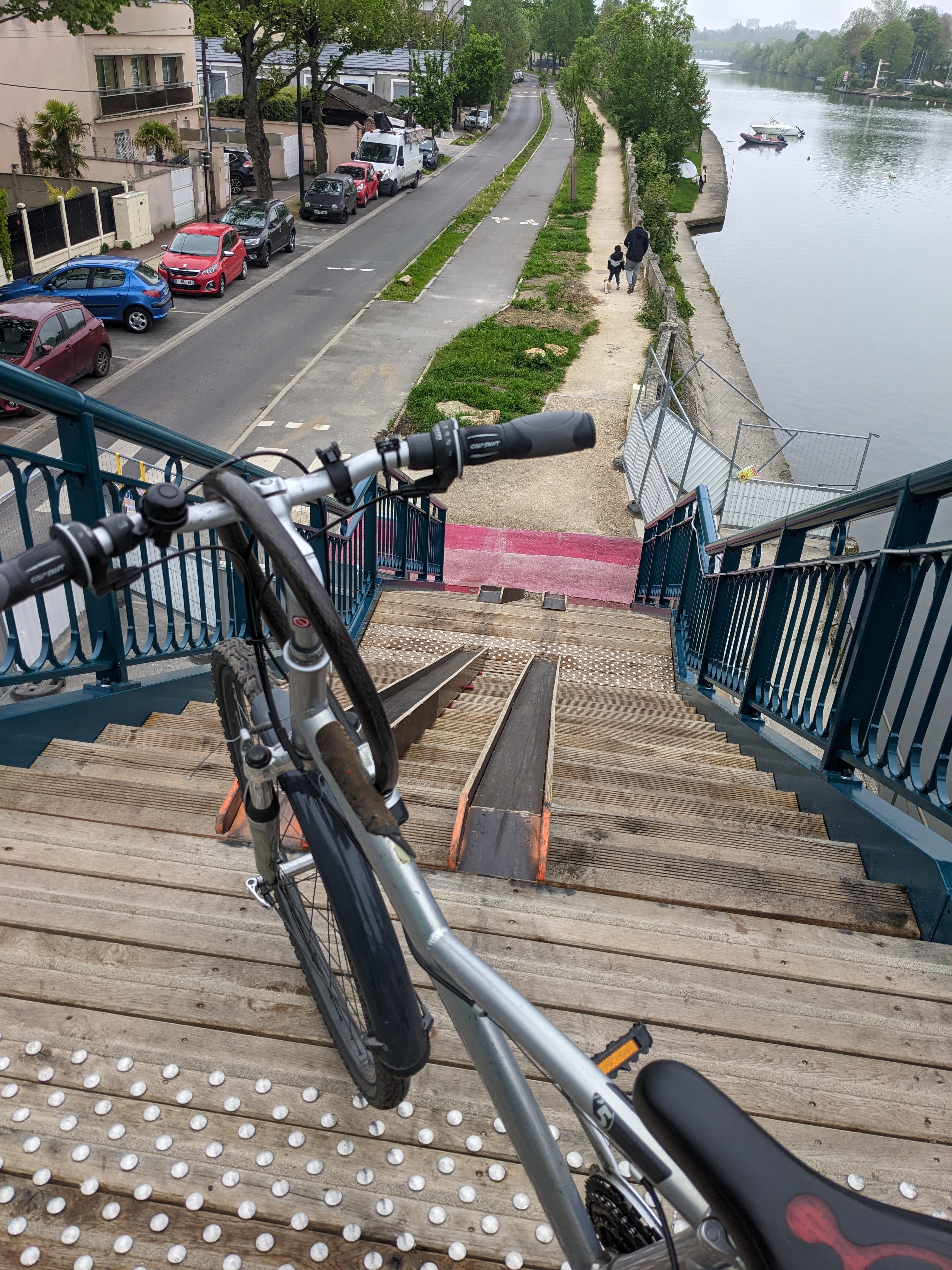 Photo : l'escalier vu d'en haut de la passerelle, le vélo tout en haut des marches, le double plan/incliné / gouttière et la Marne avec le début de la piste cyclable en contre-bas permettent d'apprécier la raideur de la pente