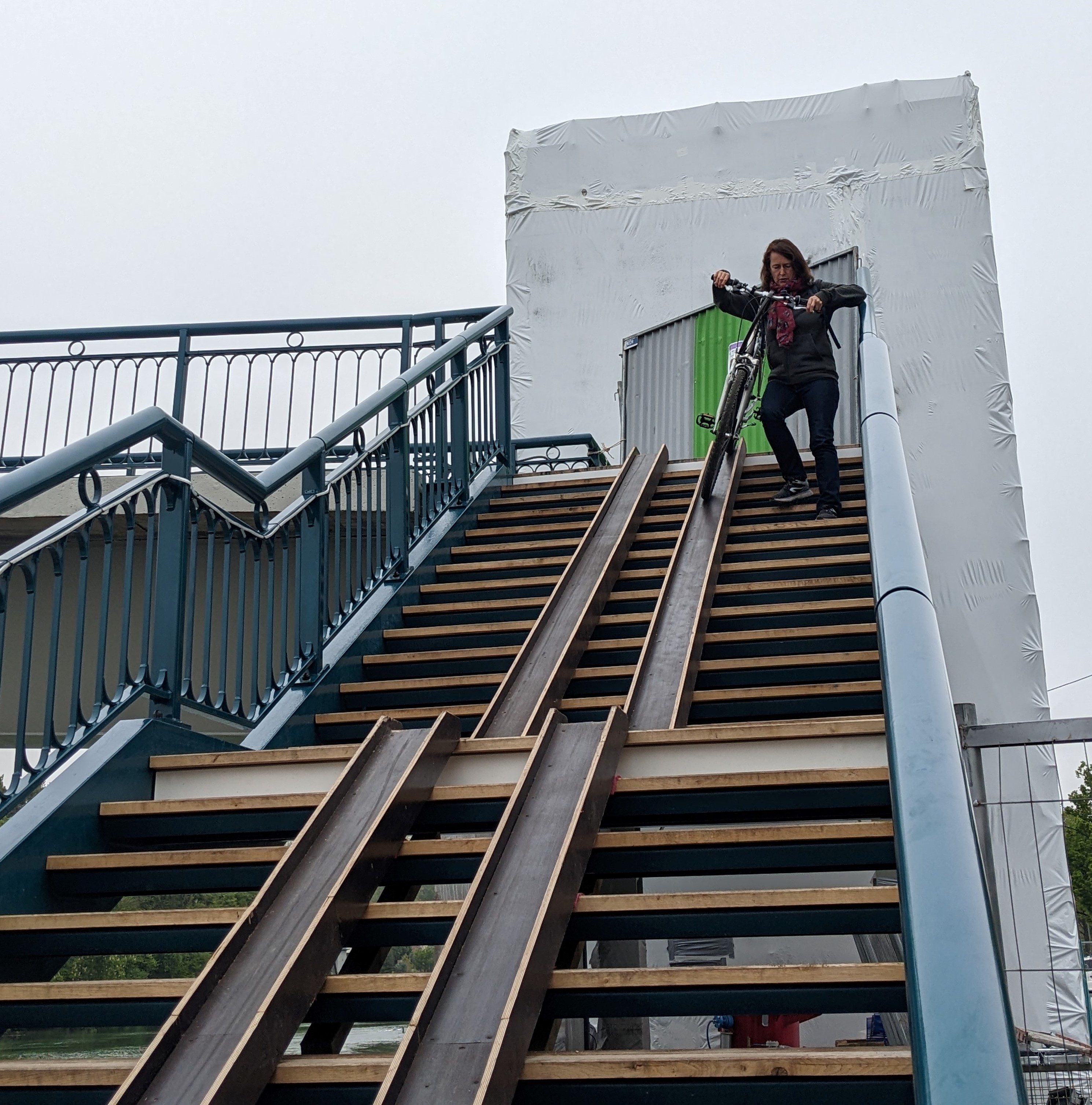 Photo : Descente de l'escalier par une cycliste tenant son vélo le long du plan incliné / gouttière, avec la cage d'ascenseur en travaux en arrière-plan. 