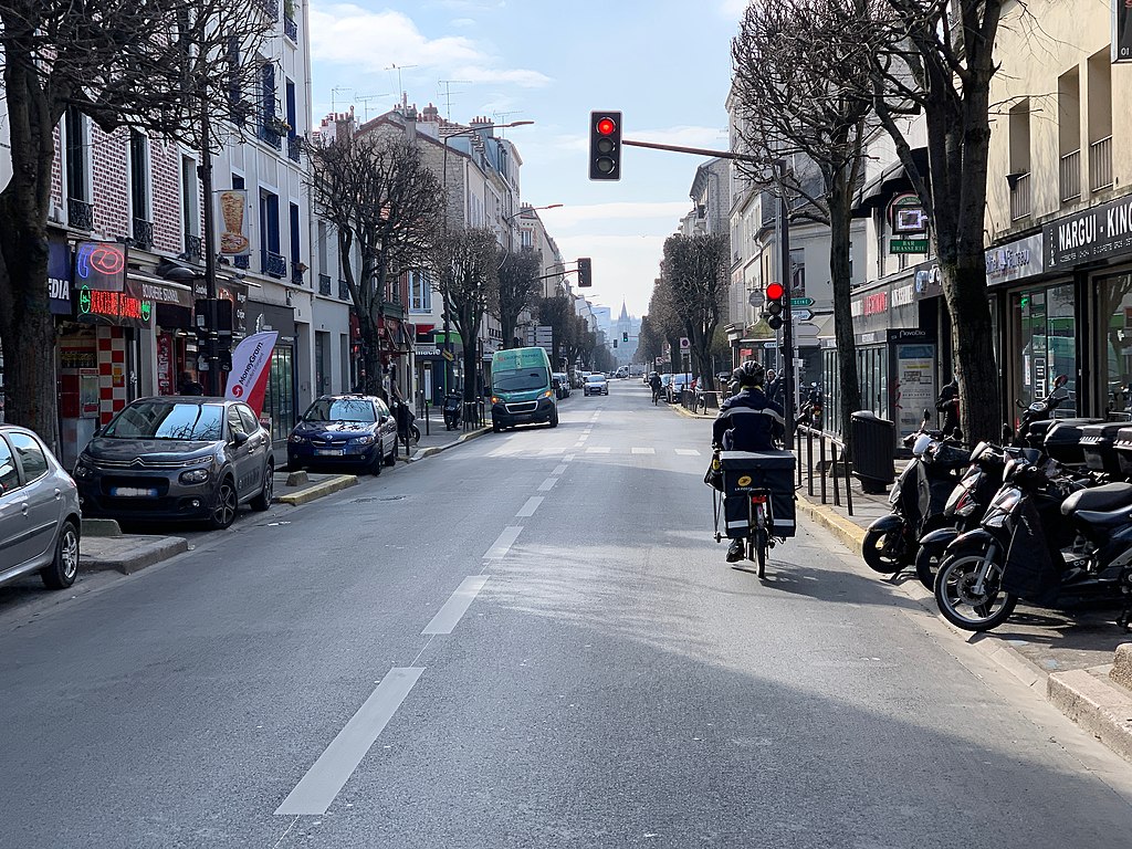 Une vue de l'avenue Paul Vaillant à Vitry-sur-Seine: deux voies du stationnement automobile et scooters est inséré de part et d'autre dans les trottoirs. Un facteur circule à vélo sur la chaussée sur la voie de droite, vu de dos. Sur la voie de gauche une camionnette verte est garée en double file, clignotants doubles apparents. 