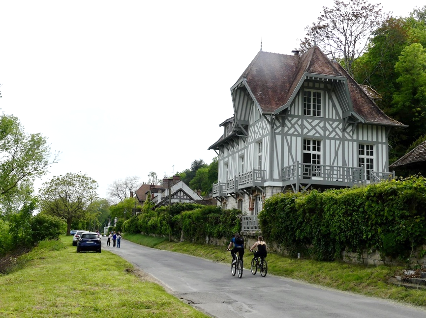 Une grande demeure bourgeoise à colombages, le long d'une route, des cyclistes et des piétons passant au pied en la regardant.