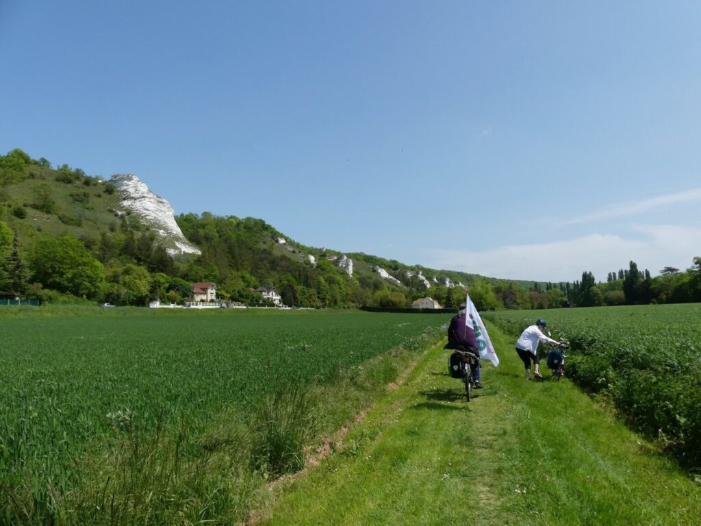 Vue d'un champ cultivé, deux cyclistes de dos sur un sentier entre deux rangées de plantations, des collines à gauche au loin