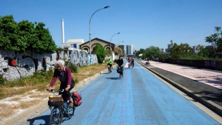 Des cyclistes roulent sur une voie peinte en bleue, des bâtiments industriels autour, proches et lointains.