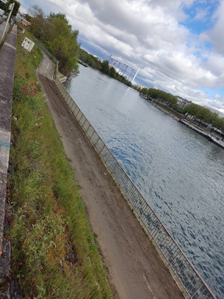 Vue d'une piste cyclable en contrebas d'une route, le long d'un fleuve, la Seine. On voit l'autre rive avec des péniches, des grues dans le lointain. La piste est couverte de boue fraîche sur une longue portion.