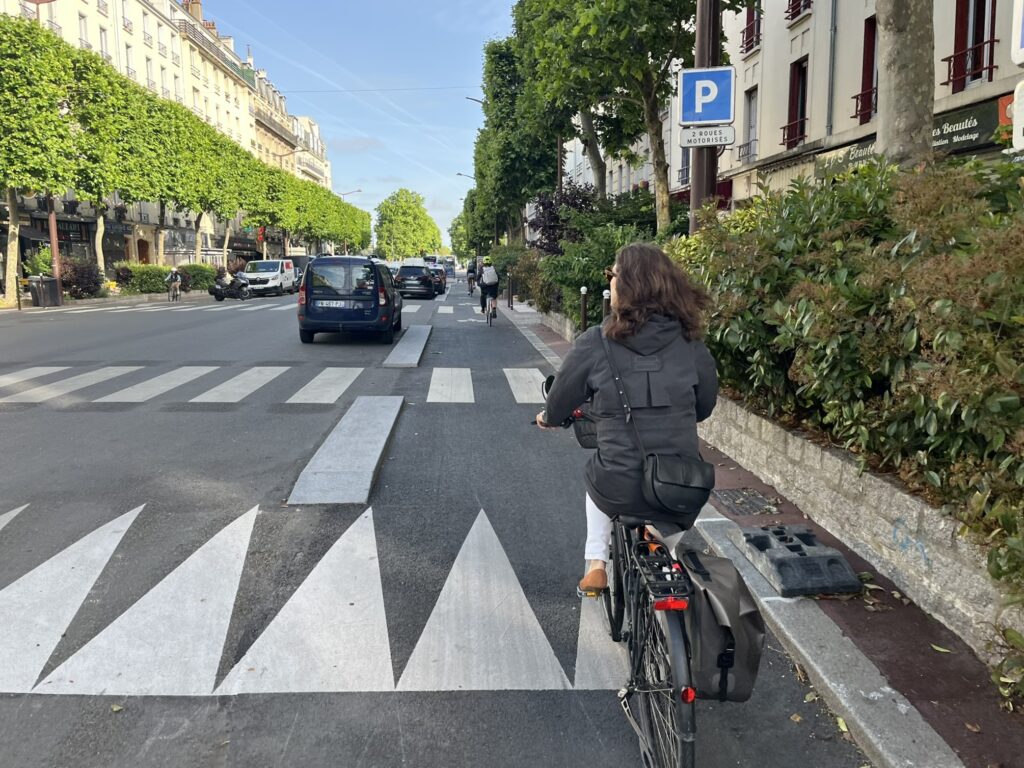 De dos une cycliste avec un imperméable sur une piste cyclable à l'approche d'un passage piéton protégé, sur un grand ralentisseur surélevé appelé plateau traversant. Devant elle plus loin deux autres cyclistes de dos.