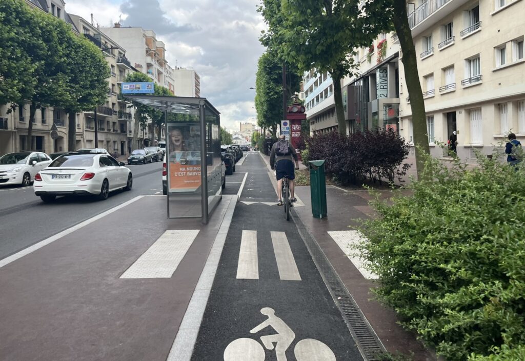 Un arrêt de bus sur la gauche, sur un îlot le long de la chaussée, une piste cyclable étroite passe à sa droite, un cycliste est de dos plus loin.