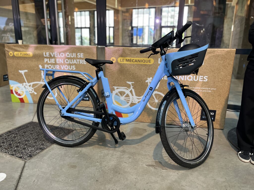 Un vélo de ville bleu simple, à cadre ouvert, avec un gros porte-bagage avant, dans une salle d'exposition.