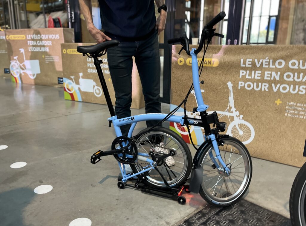Un vélo pliant à demi plié, bleu et noir. On voit les jambes d'un homme en arrière plan.