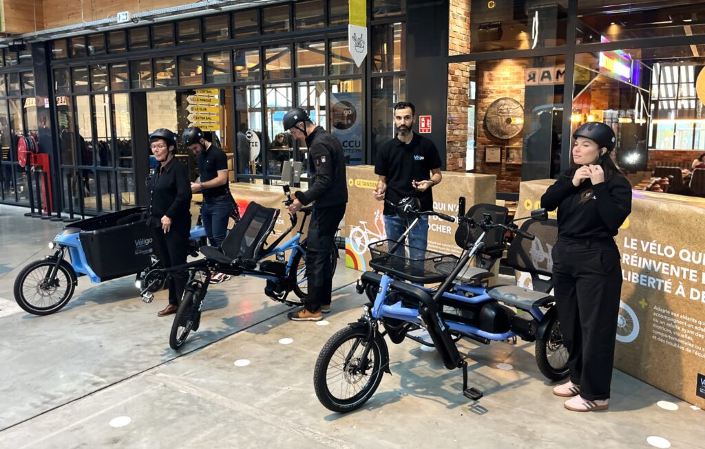 Un groupe de cinq personnes, la plupart casqués, présentent trois vélos spéciaux aux couleurs bleues et noires de la Région, dans la salle d'exposition.